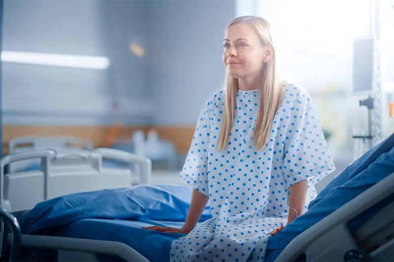 Woman sitting on a hospital bed, dressed in a hospital gown.